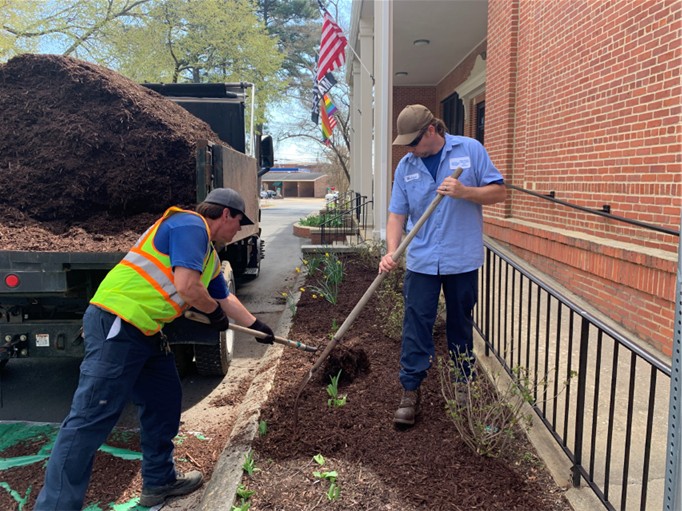Picture shows 2 Public Works employees spreading mulch in Town Hall flower beds.